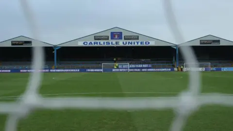 Getty Images A stand and pitch at Carlisle United ground Brunton Park, pictured through the goal net so the net string is blurry. The words Carlisle United are written on the front of one of the stand roof's peaks.