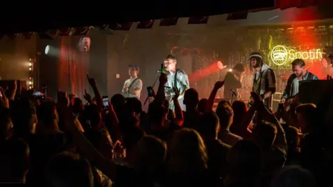 Getty Images Sam Fender e sua banda se apresentando no palco do King Street Social Club. Ele está sendo aplaudido por uma multidão, muitos dos quais mantêm os braços erguidos.