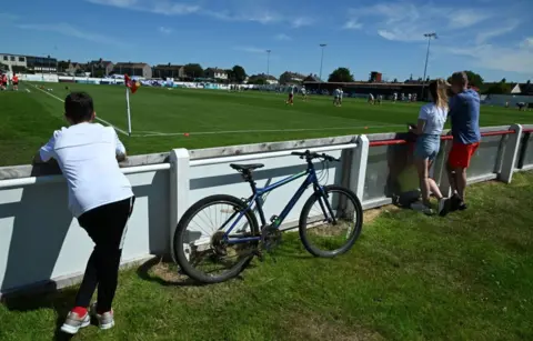 SNS Three people at the side of a football pitch, watching a game. There is a bike resting on the side of the hoardings around the pitch