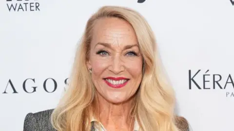 A picture of Jerry Hall, who has shoulder length blonde hair, and is wearing a white shirt and grey jacket.