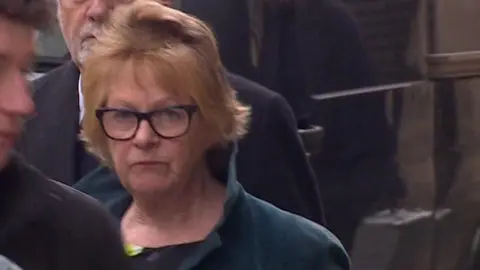 Fiona walks into court. She has short light hair, wears dark-framed glasses and wears a dark green coat and black patterned top. 