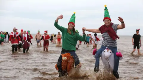 Getty Images A group of people in the sea wearing Christmas hats and costumes. At the forefront of the image are two women wearing Christmas tree hats and a bear and unicorn suit. 