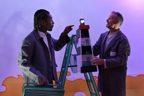 Black Country Touring Two performers can be seen on stage with a pair of step ladders behind them. One is holding a model of a lighthouse with a light on top. The other is holding a tool box. 