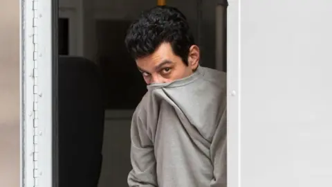 Athena Alireza Askari leaving a police van. He wears a grey sweatshirt which is pulled above his nose to cover his mouth. His head is lowered and he has dark black hair, brown eyes and his eyes are raised to the camera.