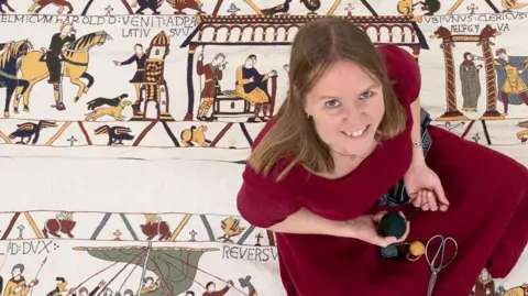 PA Media Mia Hansson, wearing a maroon dress, sits on her replica of the Bayeux Tapestry, looking up at the camera and smiling. On her lap is some thread and a pair of scissors.