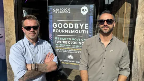 Two men wearing sunglasses and button up shirts stood in front of a shop window with a sign announcing its closure. 