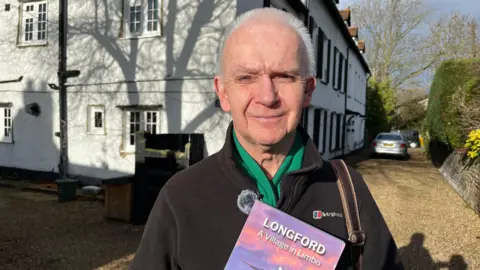 Ian Matthews, who is wearing a black fleece and green scarf holds a booklet with Longford: A Village in Limbo written upon it. He standing in front of white houses