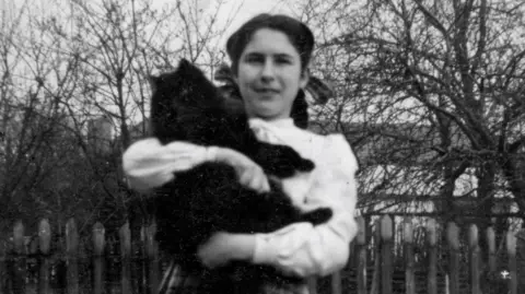 A black and white photograph of a dark-haired girl wearing a white blouse and holding a large black cat.