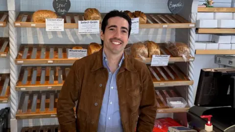 Carlos Huerta Carlos Huerta wearing a blue shirt and brown jackets smiles at the camera. Loaves of bread are on the display shelves behind him. 