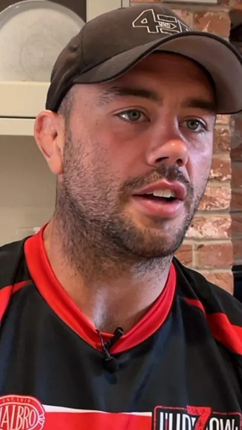 Man with a cap and rugby shirt talking to someone off camera. 