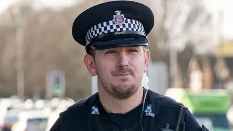 Paul Hogben wearing a black police hat and the black uniform with a walkie-talkie attached to his chest. He is walking by a road with queuing traffic.