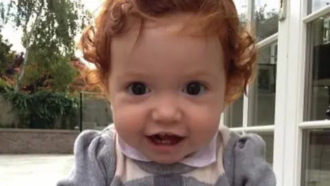 A toddler with ginger curly hair grins at the camera while sat in a garden. She is wearing a grey jumper with red hearts on