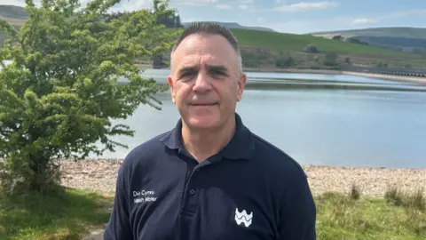 Man wearing a Welsh Water navy polo shirt looking at the camera with Crai Reservoir in the background