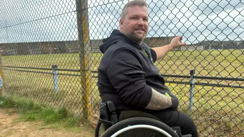 Matt Precey/BBC Man in a wheelchair looking at the camera. He is next to a chain link fence which he is holding on to. He has a bead and is wearing a dark hoodie.