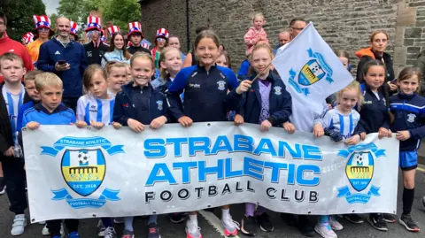 A young football team pose for a photograph during a parade, holding up a banner of their club Strabane Athletic