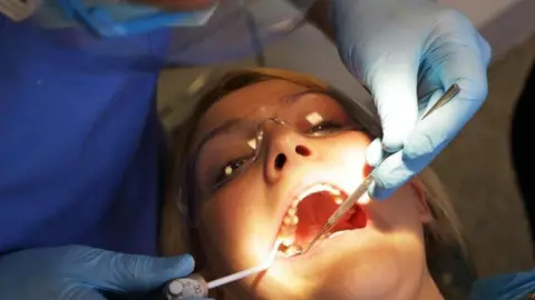 A woman wearing glasses and with her mouth open is looking at the camera as dentals objects are in her mouth. Someone wearing light blue gloves near her face is holding the objects.