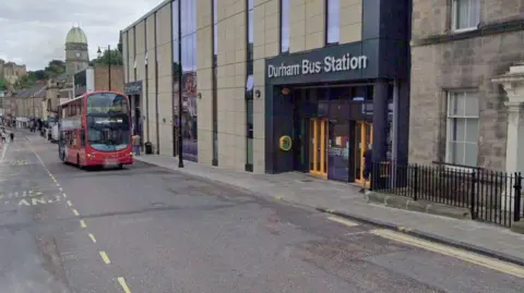 A Google Streetview screenshot of North Road outside the bus station in Durham. A double decker bus is travelling down the road on the right-hand side. The left side of the road has road markings for a bus stand all the way along it. 