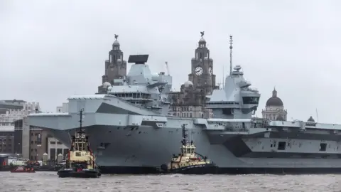 The HMS Prince of Wales in the River Mersey, with the Liver Bird buildings in the background.