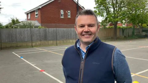 James Grant/BBC A man in a blue jumper and a navy gilet stands in a playground smiling at the camera. 