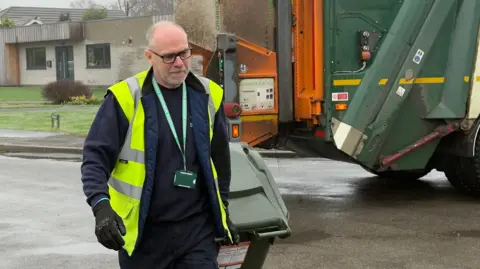 Keith Sayers moving a wheelie bin on a street, with a bin lorry in the background