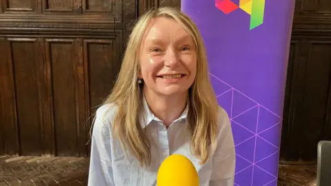A woman with long dark blonde hair is wearing a white blouse and smiling at the camera.