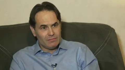 A middle-aged man wearing a light blue button-up shirt sits on a grey sofa. He has a small lapel microphone attached to his shirt and his facial expression is serious. 