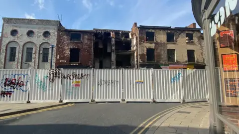 Grace Wood/BBC A picture showing a row of derelict buildings on a street with safety barriers cordoning them off. The barriers have graffiti writing on them. 