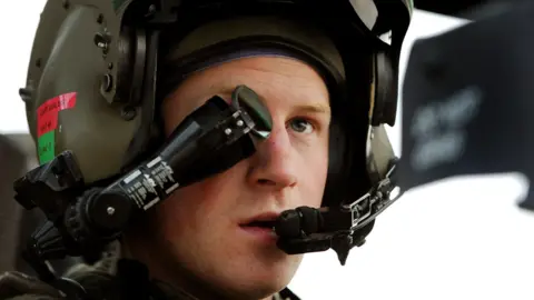 Prince Harry in 2012 wearing a green helmet attached to an eyepiece that is over one eye and a mouthpiece in front of his mouth