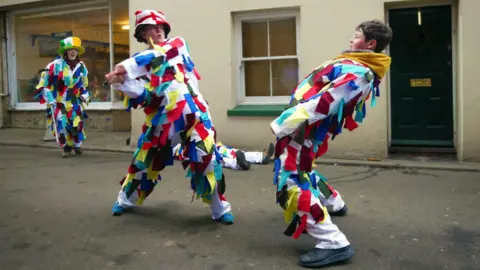 Paul Hardman A young girl and a boy wearing white outfits covered in multi-coloured ribbons having a mock sword fight in a village street. The girl, who is wearing a hat with the English flag on it, is swinging the sword and the boy is ducking backwards. A third person in similar dress, with a hat with the Irish flag on it, cheers them on in the background.