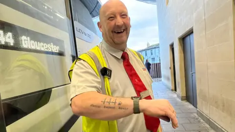 A man is showing a tattoo on his forearm which says "444. 4better. 4worse. 4ever". He is wearing a smart watch too, and a short sleeve shirt and high visibility jacket. He is smiling and laughing joyfully at a camera. He is stood next to a large coach, which says 444 Gloucester on an electronic screen.