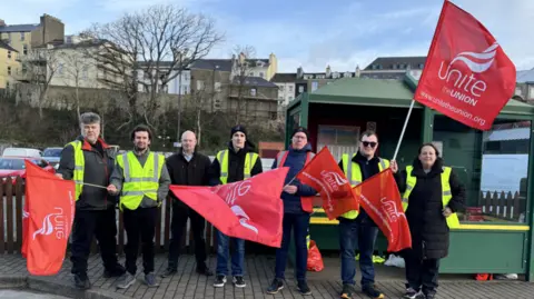 A line of men and a woman are standing on a pavement with a green bus stop to the right behind them. They are wearing high vis jackets and waving red flags that read Unite the Union.