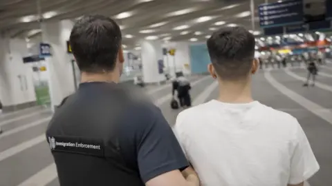 Home Office An officer wearing an Immigration Enforcement best his arm hooked under the arm of a man, taking him away. The man looks young but we can only see the backs of their heads. They are in a train station.