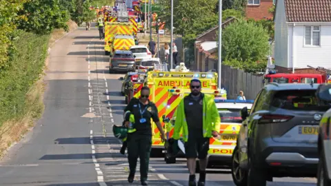 PA Media A large emergency personnel presence on a road parallel to the railway line. There are police vehicles, fire appliances and ambulances parked on the side of the road. Some emergency personnel are walking up the road or standing outside vehicles. Two ambulance workers are walking towards the camera, one wearing a hi-vis fluorescent jacket and the other carrying items in her arms. 