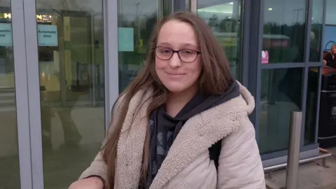 A young woman wearing a black hoodie and cream coloured coat. She has brown hair and is wearing glasses. 