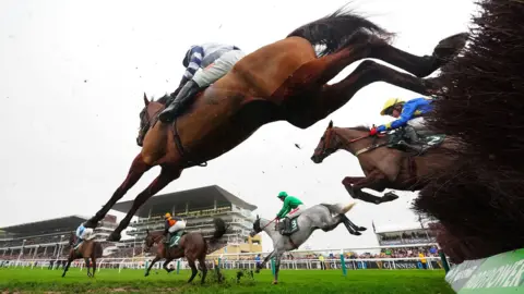 PA An upwards view of horses jumping over a jump at Cheltenham race course taken from the ground behind the jump. There are three horses jumping over with two already clear.