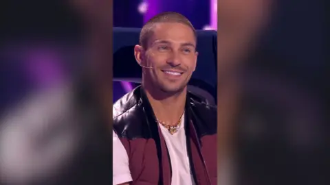 A man, Joey Essex, smiles, showing off his bright white teeth. He has a shaved head and wears a body warmer