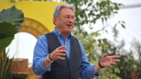 RHS Malvern A man in a blue shirt and waistcoat who is talking and has his arms raised. Behind him is a yellow arch and green trees and plants. 