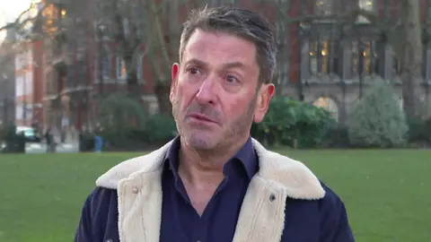 Sean Connaughton, wearing a fleece jacket, is interviewed in a park with a building in the background