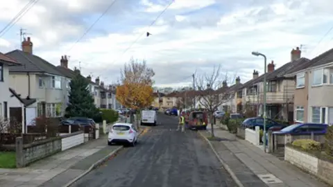 A white car and a van are parked on the pavements on Keble Drive by trees and rows of houses