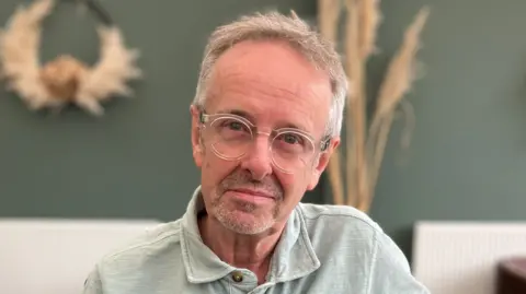 A man with short grey hair looks directly into the camera. He is wearing a light green polo shirt and clear plastic rimmed glasses. The background is out of focus but there is a green wall with dried flower decorations and white radiators behind him. 