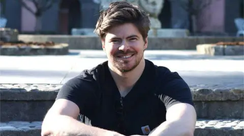 Wolverhampton Literature Festival A man in a black t shirt, sat on steps. He has a brown beard and hair. He is smiling at the camera