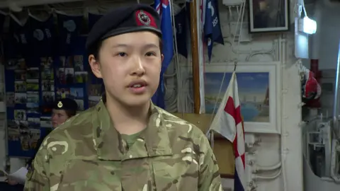 A teenage girl in a military uniforms stands in front of a flagpole.