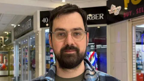 Andy Blair is pictured in a shopping centre. He has short dark hair, a dark beard and is wearing silver framed glasses and a blue checked shirt. 
