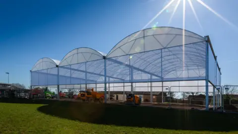 Contributed Three padel courts being constructed on a sunny day. Three large domes have been constructed above the courts. Construction equipment can be seen below the domes. 