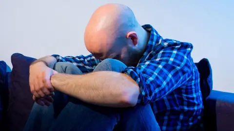 A bald man wearing a blue chequered shirt sits in a crouched position on a sofa with his face sunk towards his knees and his arms wrapped around his legs.