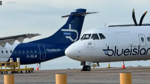 Two Blue Islands planes on the tarmac at Guernsey Airport. To the right of the image is the nose and front section of one aircraft. It is white, with navy blue writing saying 'blue islands'. A propeller is visible on the right of the image. To the left of the image is the tail of another blue islands plane. It is navy blue, and has a logo in the shape of a 'b', which is white. In front of the plane is a piece of yellow equipment with wheels.