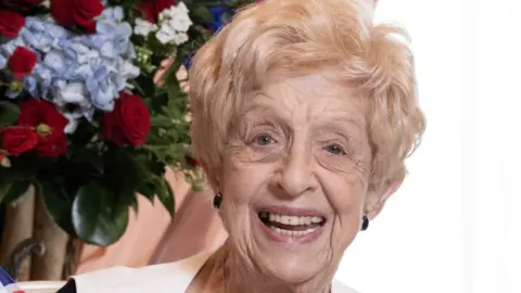 Ruth June Bourne has short blonde hair and is smiling in front of a bouquet of roses.