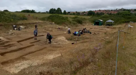 An archaeology trench dug into the soil with people working in it. There are two large tents, a field and some houses in the background. 