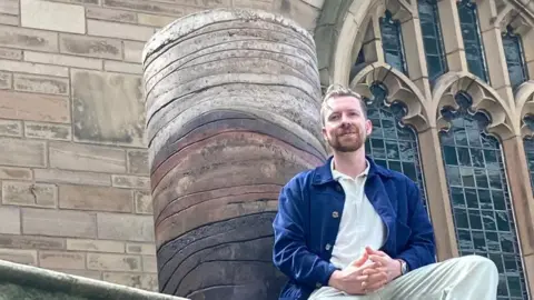 Benjamin Stephenson is wearing beige trousers and a blue jacket over a white shirt. He is sat at the base of his sculpture which is a cylinder showing layers of brown compacted soil and clay.