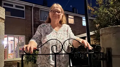 Kevin Shoesmith/BBC A bespectacled woman with greying light brown hair rests her hands on a wrought iron gate in front of her home. The photo is taken at night, with light coming from the living room behind her. 